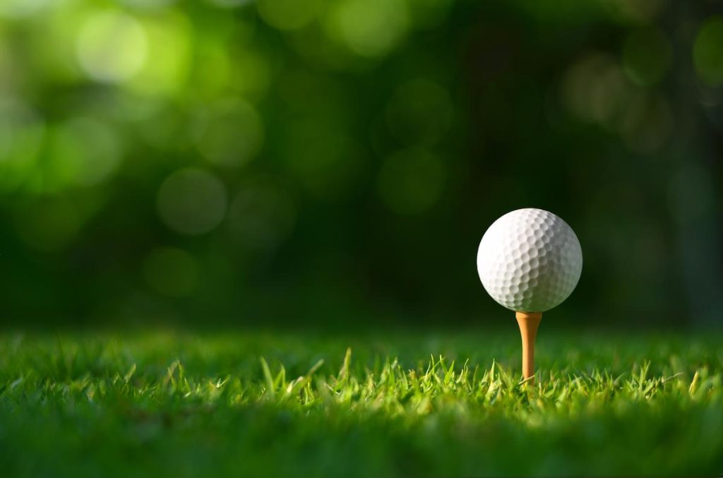 A close-up, low-angle shot of a white golf ball resting on a wooden tee in the rough of a golf course. The ball is clearly visible against the bright green grass in the foreground. The background is a deeply blurred bokeh of dark green foliage and sunlight filtering through the trees, creating a soft, warm atmosphere.