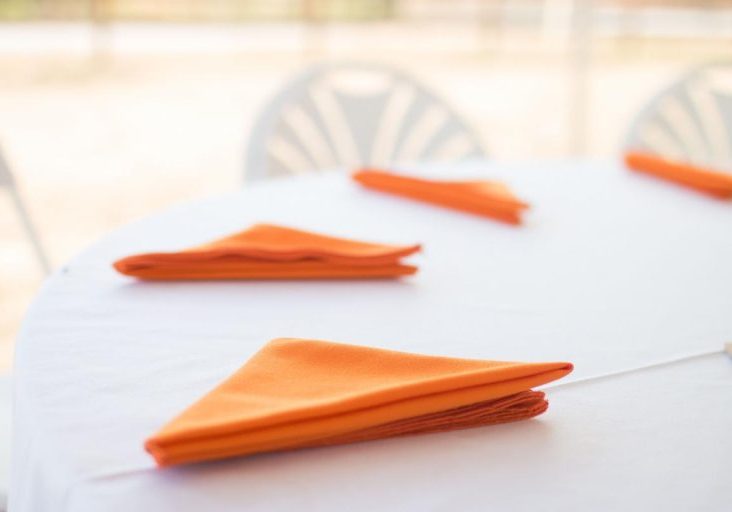 A close-up, high-key image of a round table covered with a crisp white tablecloth, set up for an event. Several brightly folded orange cloth napkins are placed diagonally on the table, indicating place settings. In the blurred background, the white backs of folding chairs and a bright, indistinct outdoor setting are visible, suggesting an event like a fundraiser or reception.